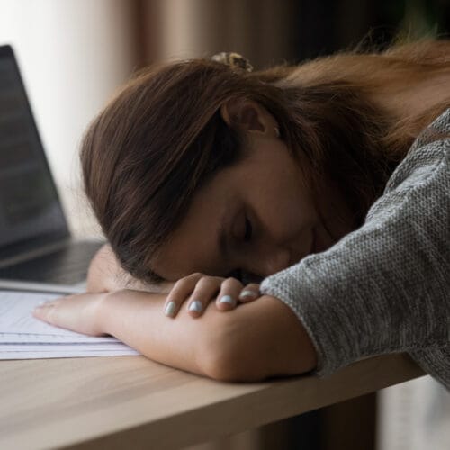 A sleeping woman rests her head on her arms on a desk with a laptop, papers, and a cup of coffee nearby.