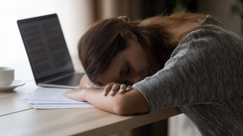 A sleeping woman rests her head on her arms on a desk with a laptop, papers, and a cup of coffee nearby.