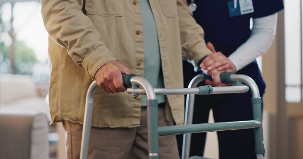 Close-up of an elderly person using a walker with support from a caregiver placing a hand on the walker for assistance.
