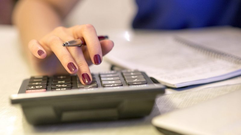 A close-up of a woman's hand using a calculator while holding a pen, with a notebook visible in the background.