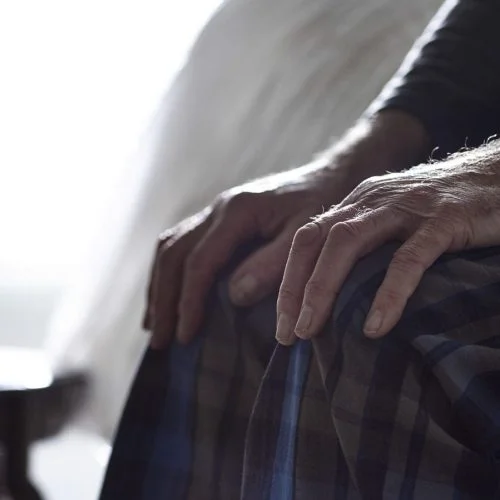 Close-up of an elderly person's hands resting on their lap while sitting on the edge of a bed.
