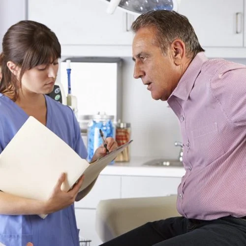 Middle-aged man holding his lower back in pain while consulting with a doctor and nurse in a medical office. The nurse is taking notes.