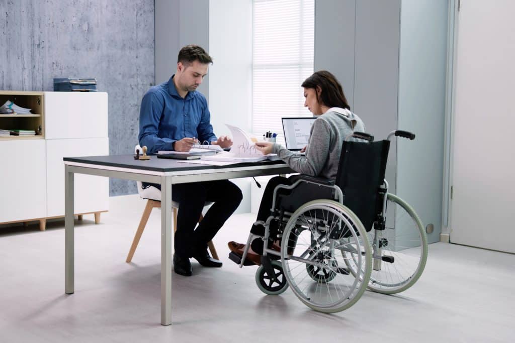 A woman in a wheelchair looks through a binder full of papers with her attorney. A laptop is also on the desk and there is a filing cabinet in the background.